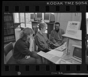A photograph of 3 people sitting at a table in front of a computer.