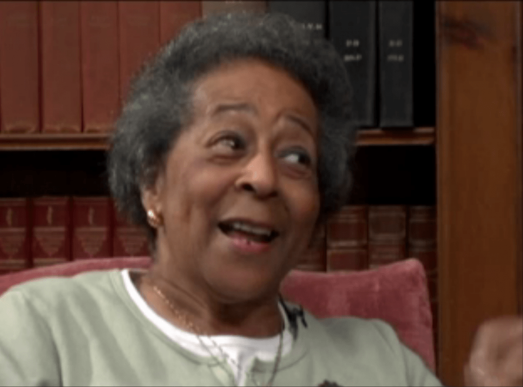 An African-American woman with grey hair smiles as she looks off-camera towards her interviewer. She is wearing a green sweater and sits in an armchair. Bookshelves fill the wall behind her.